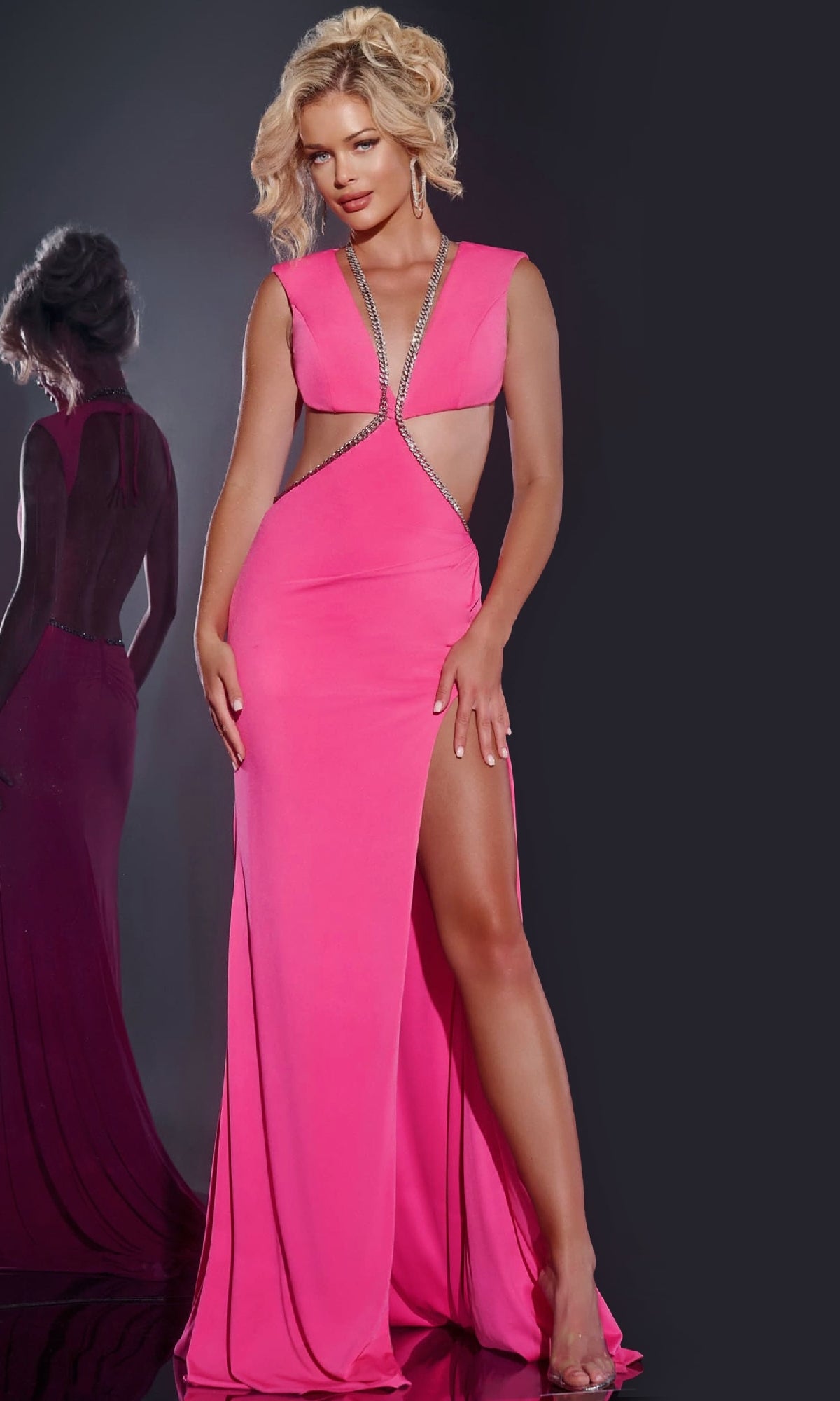 A blonde woman wears the Jovani 42675 long prom dress in pink, featuring cut-out details and a thigh-high slit, standing before a dark background with her reflection visible.